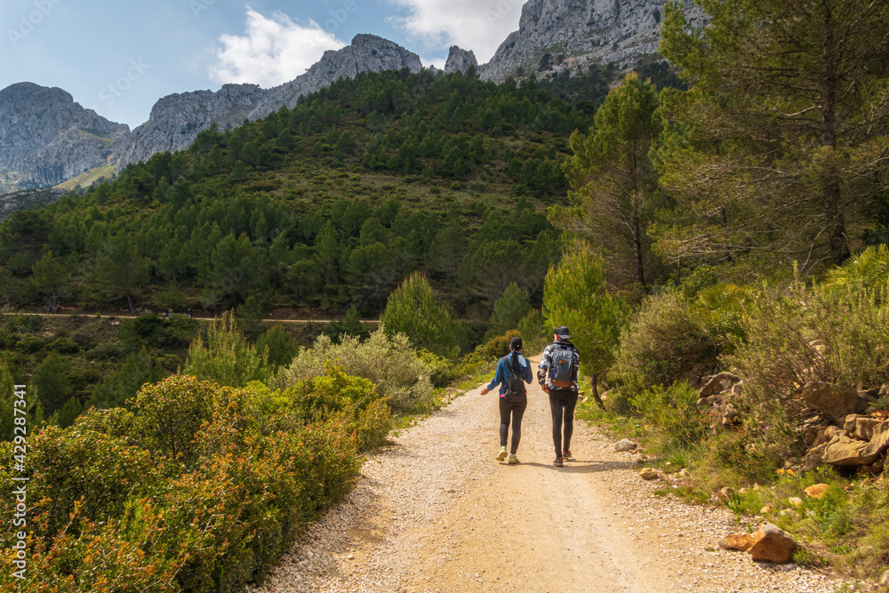 People walking along a path, near the Sierra de Bernia mountains, on a spring day, with cloudy skies.