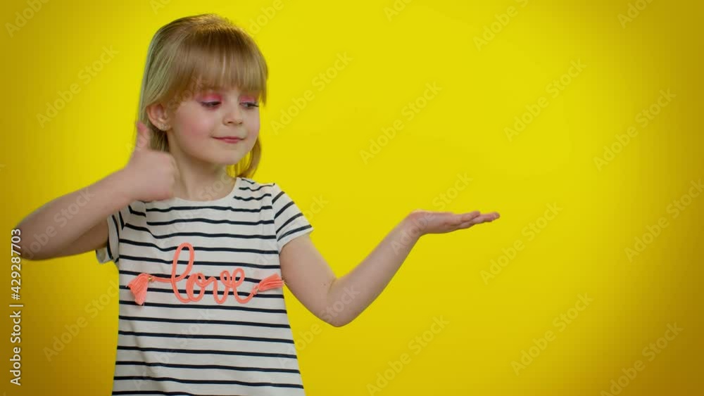 Funny playful blonde kid child showing thumbs up and pointing empty place, advertising area for commercial text, copy space for goods promotion on yellow studio background. Cute teenager children girl Funny playful blonde kid child showing thumbs up and pointing empty place, advertising area for commercial text, copy space for goods promotion on yellow studio background. Cute teenager children girl