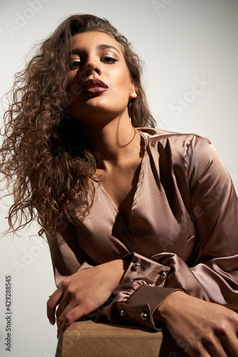 Fashion portrait beautiful brunette woman with curly hair in stylish in brown satin dress. Concept of model posing in trendy accessories and clothing in professional studio. 