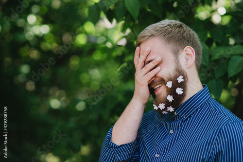 Young smiling caucasian man with lilac flowers in his beard having fun and posing for a photo.