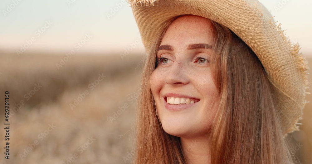 Close up of pretty happy young woman standing in golden field and putting on hat in good mood. Beautiful cheerful girl smiling and posing outdoor in countryside with smile on face. Portrait concept