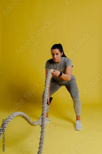 Athletic young woman doing some crossfit exercises with a rope indoor on yellow background.