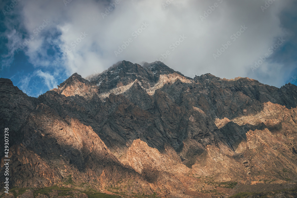 Mountain landscape with rocks in golden sunlight and low clouds. Awesome rocky wall with sharp top in gold sunshine in overcast weather. Atmospheric scenery with high rocky mountain pinnacle in clouds