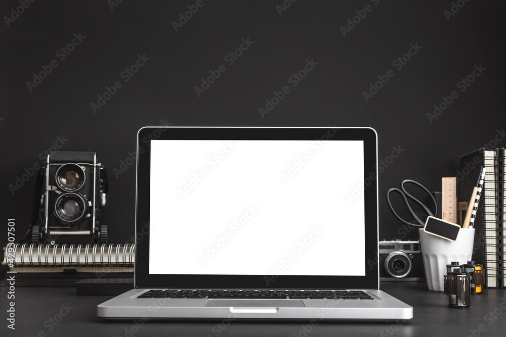 Creative desk with a laptop screen, camera, desk objects, office supplies, books and black wall. Mock-up.	
