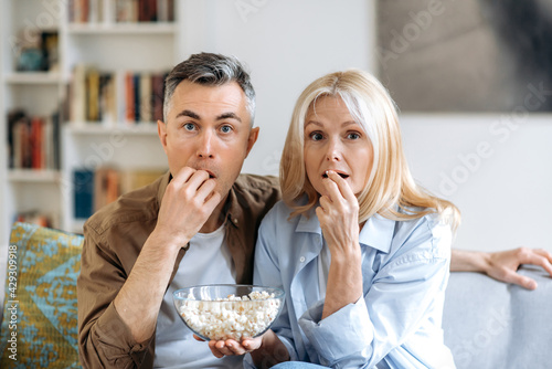 Middle aged shocked caucasian family couple sitting at home on sofa in casual clothes  watching tv show or exciting movie  eating popcorn  spending weekend together at home