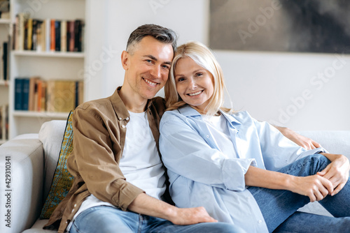 Portrait of a pleasant happy mature caucasian married couple relaxing on sofa in living room at home, spending time together, dressed in casual stylish clothes, smiling at camera, happy together