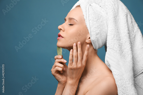 Beautiful young woman doing facial massage with gua sha tool on blue background. Space for text photo