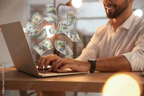 Making money online. Closeup view of man using laptop at table and flying dollars photo