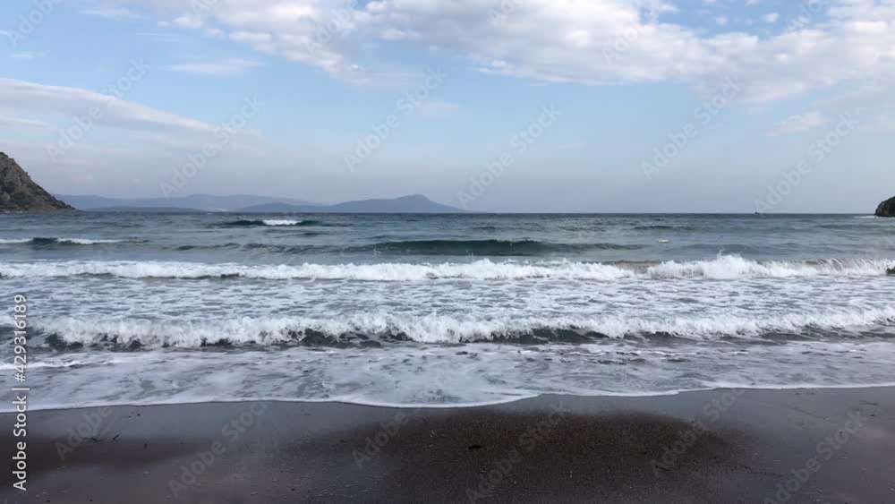 Many white foamed waves approaching the beach. Grey blue sky with clouds. Dark sea. Black sandy coast. Islands at horizon. Natural ocean background with copy space. 4K resolution video Many white foamed waves approaching the beach. Grey blue sky with clouds. Dark sea. Black sandy coast. Islands at horizon. Natural ocean background with copy space. 4K resolution video