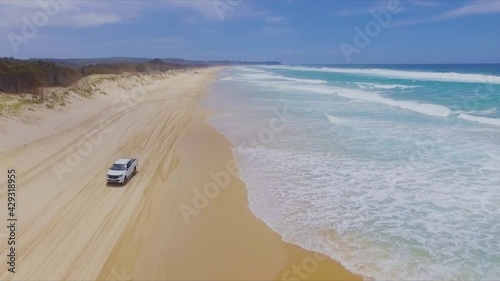 Low aerial drone shot travelling North up Main Beach on Queensland's North Stradbroke Island, as a white 4x4 four wheel drive pickup truck vehicle drives South on the beach below