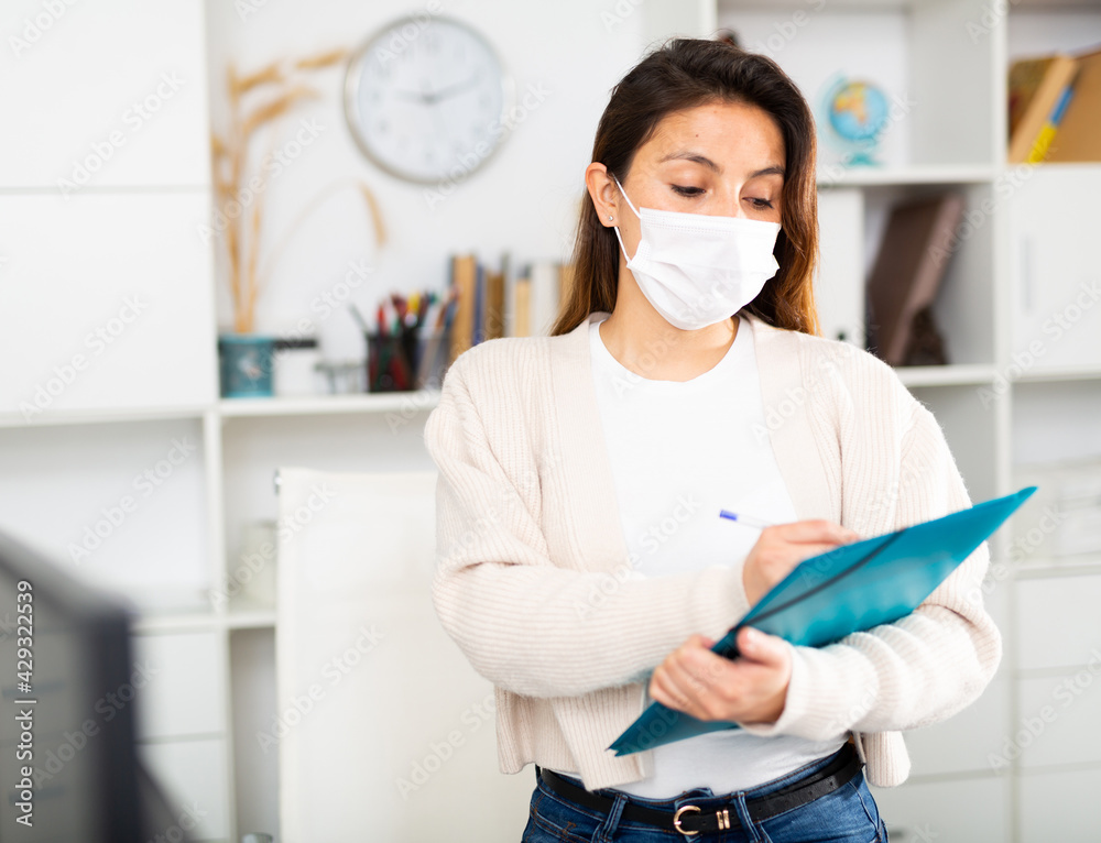 Confident girl manager stands in a protective mask during the pandemic next to the workplace in the office and fills ..out documents on a folder