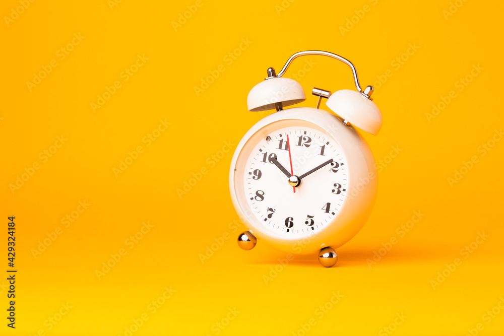 Small white alarm clock, black numbers, set the time placed on a table. Clock on isolated yellow background. Small white alarm clock, black numbers, set the time placed on a table. Clock on isolated yellow background.