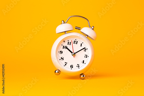 Small white alarm clock, black numbers, set the time placed on a table. Clock on isolated yellow background.