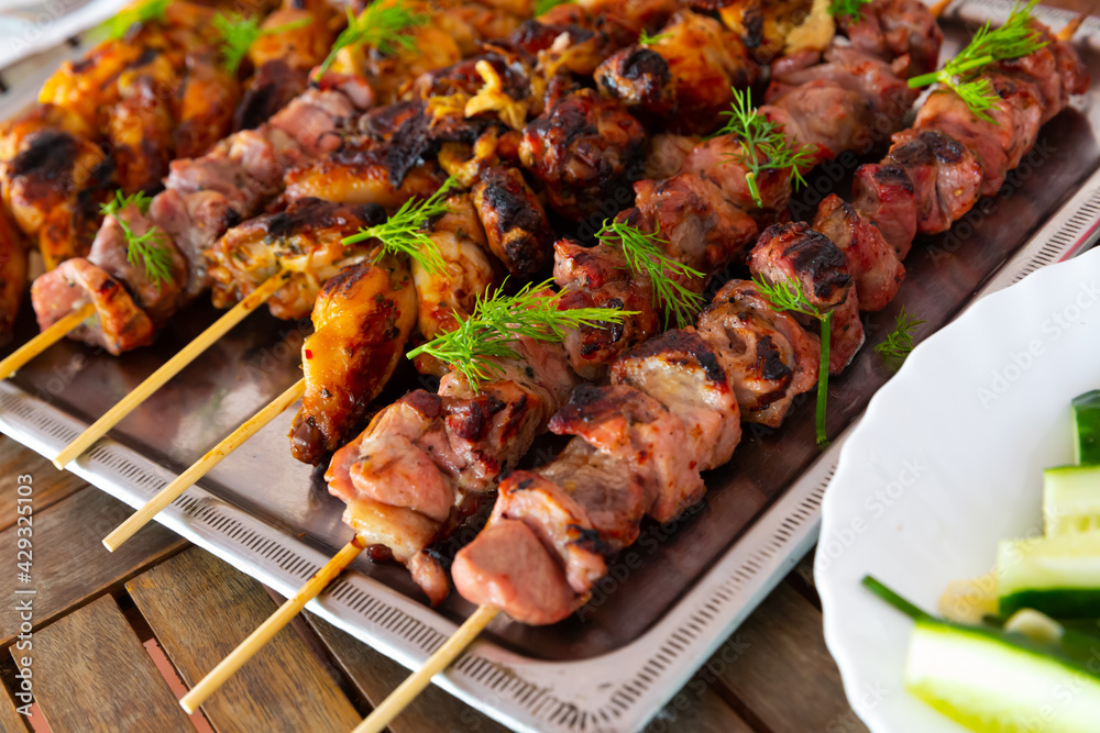 Tasty grilled chicken and pork skewers garnished with fresh greens served with cucumbers on metal tray