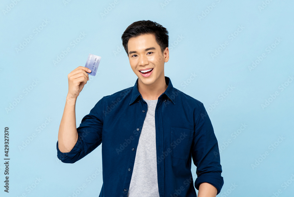 Waist up portrait of happy smiling young handsome Asian man holding credit card in isolated light blue background Waist up portrait of happy smiling young handsome Asian man holding credit card in isolated light blue background
