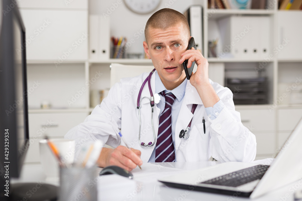Doctor with stethoscope in white modern office working on laptop and talking on phone