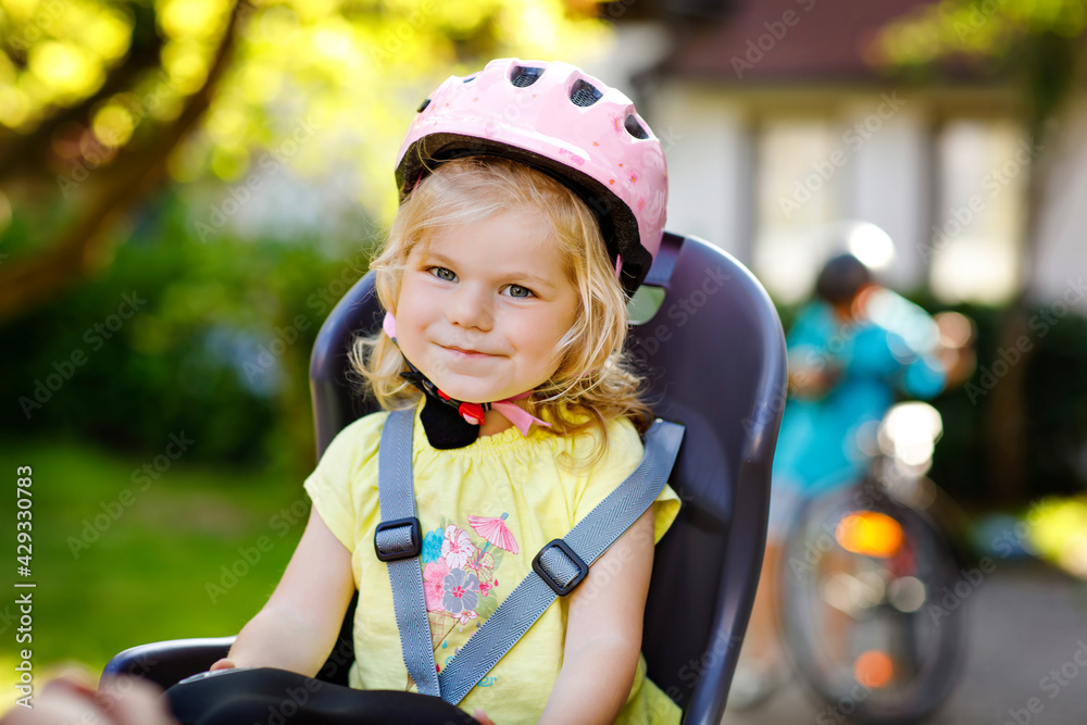 Portrait of little toddler girl with security helmet on the head sitting in bike seat of parents. Boy on bicycle on background. Safe and child protection concept. Family and weekend activity trip. Portrait of little toddler girl with security helmet on the head sitting in bike seat of parents. Boy on bicycle on background. Safe and child protection concept. Family and weekend activity trip.