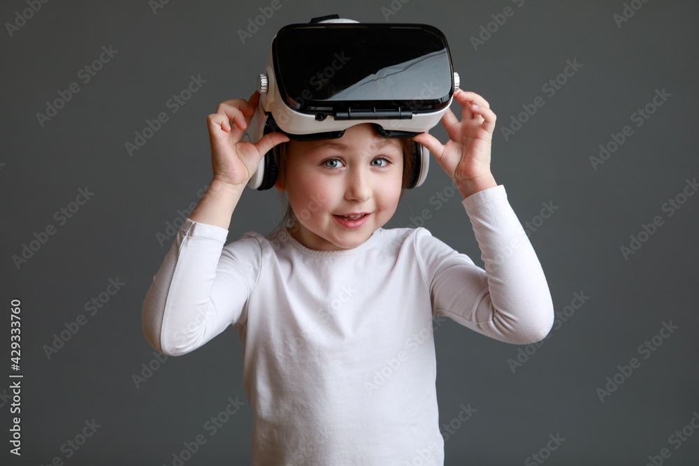 Smiling little kid girl in white long sleeve shirt isolated on grey background wearing VR glasses Smiling little kid girl in white long sleeve shirt isolated on grey background wearing VR glasses