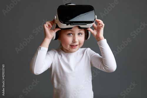Smiling little kid girl in white long sleeve shirt isolated on grey background wearing VR glasses