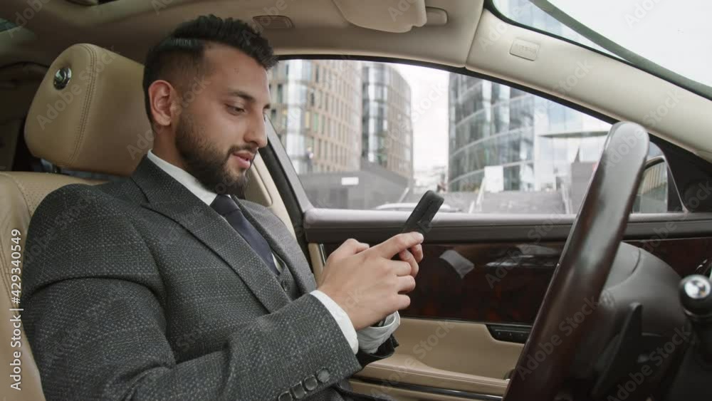 Low angle medium close-up young professional Mixed-Race male driver wearing formal suit, sitting in front seat in automobile, using smartphone, then making call Low angle medium close-up young professional Mixed-Race male driver wearing formal suit, sitting in front seat in automobile, using smartphone, then making call