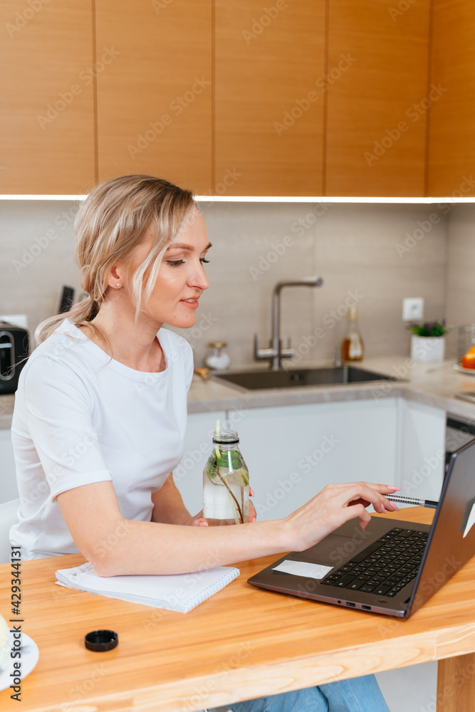 Blonde girl with laptop drinking majito lemonade, doing homework in a bright kitchen. Education and business concept. remote work and freelance work. Business lady. vertical photo Blonde girl with laptop drinking majito lemonade, doing homework in a bright kitchen. Education and business concept. remote work and freelance work. Business lady. vertical photo