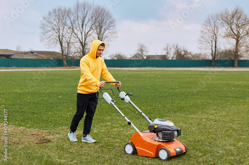 Young attractive man trimming grass with cutter, young adult male wearing yellow hoodie and black trousers mowning lawn, pushing special equipment for taking care of field.