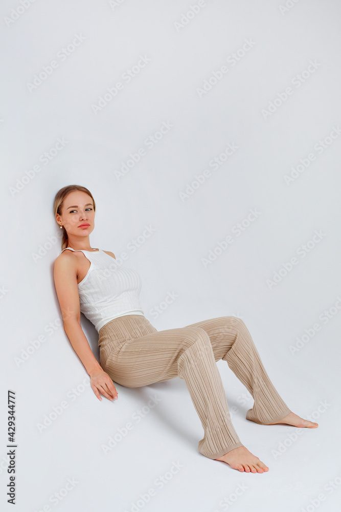 A young beautiful blonde in a fashionable white T-shirt and beige knitted trousers sits on a cyclorama on a white background in the studio.
