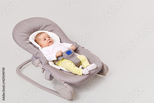 Tableau sur toile Side view of charming new born girl lying in bounce child bouncer chair, little baby in tiny clothes shirt and yellow pants infant lying in rocking chair, isolated over white background