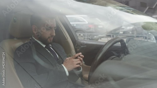 Medium outside car through front window view of young Mixed-Race businessman wearing formal clothing, sitting behind wheel, having work call on mobile phone photo