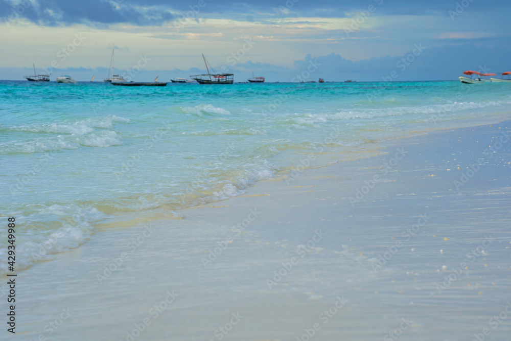 Seascape with white yellow sandy beach, small waves of bright blue indian ocean with boats silhouettes at the horizon and cloudy sky in the early morning in South Africa Tanzania Zanzibar island