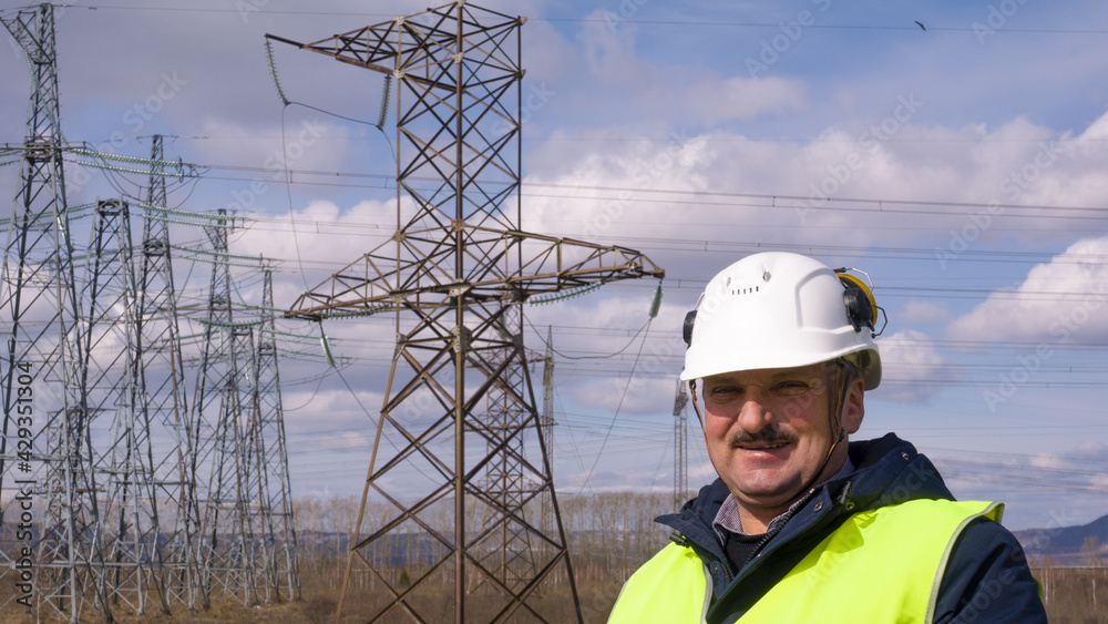Smiling middle-aged man wearing protective helmet on his head and reflective safety vest near high-voltage power lines makes a detour around the equipment. Electrical engineer, electrician