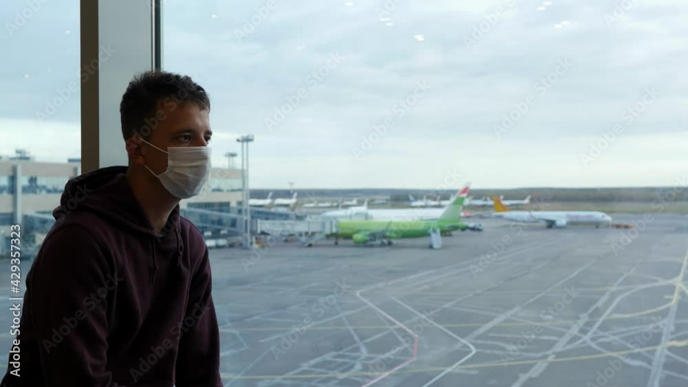 Masked man in an airport terminal sits by a masked window and looks at an open-air plane park at the airport. Male passenger traveling by plane during the covid19 pandemic. Masked man in an airport terminal sits by a masked window and looks at an open-air plane park at the airport. Male passenger traveling by plane during the covid19 pandemic.