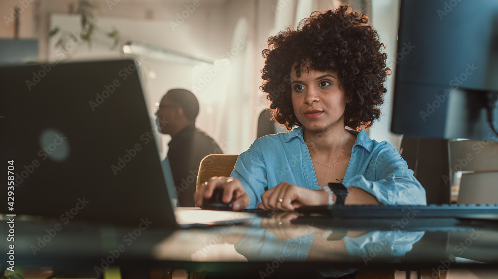 Creative Office: Young Black Woman Sitting at Her Desk Working on Computer. Charmingly Authentic Software Developer, Social Media Marketing Specialist Creating Content, Using Devices. Wide Shot Creative Office: Young Black Woman Sitting at Her Desk Working on Computer. Charmingly Authentic Software Developer, Social Media Marketing Specialist Creating Content, Using Devices. Wide Shot