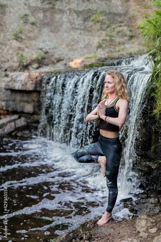 Blonde woman in sportswear practicing yoga at the forest riverside near the waterfall. Fitness girl doing a balance exercise  standing on one leg with hands clasped with in namaste mudra sign