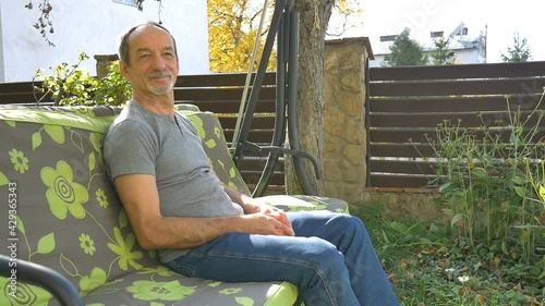 Senior man is sitting on a swing outdoor during summer sunny day on playground in the garden and thinking about something