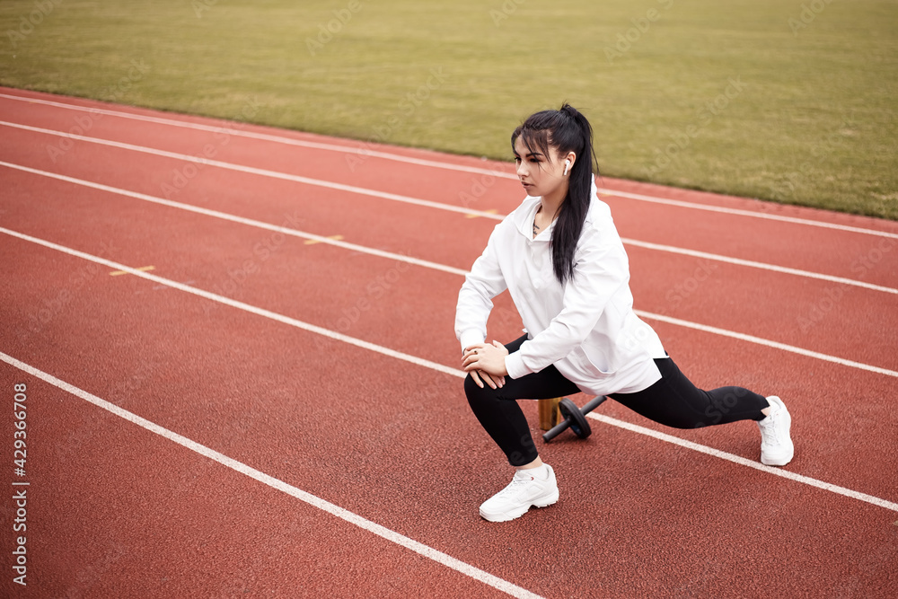 Girl stretches, prepares her body and muscles for a productive fitness workout. Flexible female dressed sports suit sporty model on the city stadium. Image with copy space. Single leg squats
