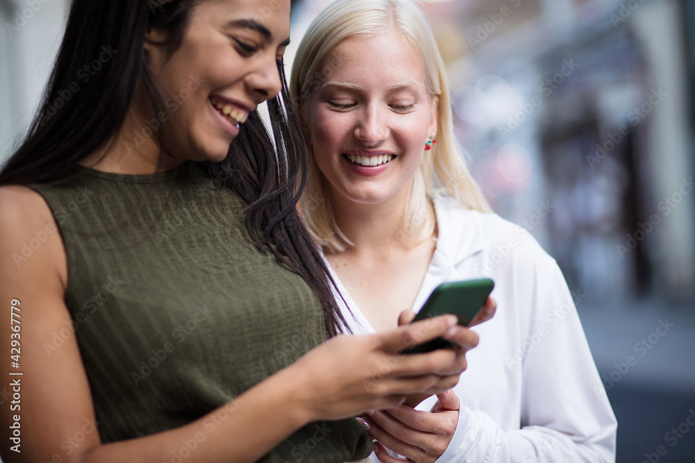 Two women on street using phone together. Focus is on blond woman. Two women on street using phone together. Focus is on blond woman.