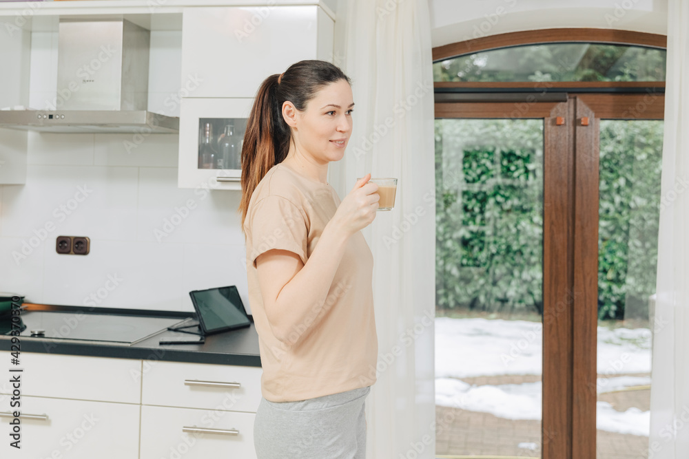 Young woman in casual clothes is drinking cappuccino or caffee in her kitchen. Housewife is happy in the kitchen in the morning Young woman in casual clothes is drinking cappuccino or caffee in her kitchen. Housewife is happy in the kitchen in the morning