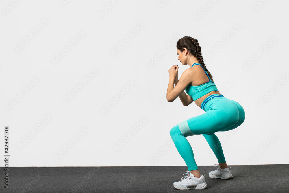 Rear view shot of a sporty woman dressed in sportswear. Female model doing squats and stretching exercise for buttocks on white background