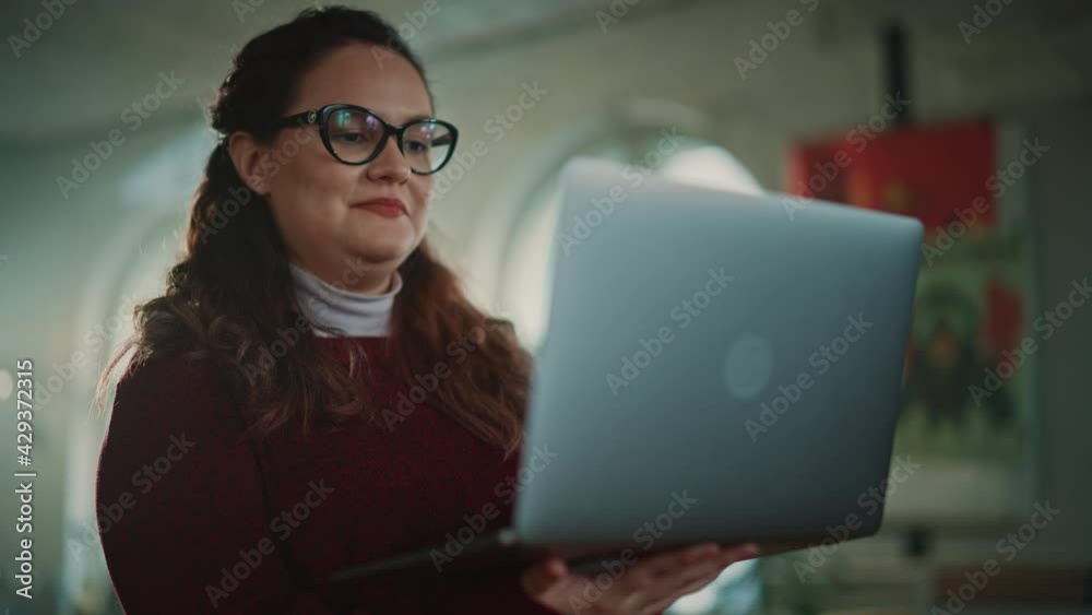 Creative Office: Young Hispanic Woman Holding Laptop Computer and Working. Charmingly Authentic Smiling Businesswoman with Stylish Glasses Creates Social Media Marketing Product. Medium Shot Creative Office: Young Hispanic Woman Holding Laptop Computer and Working. Charmingly Authentic Smiling Businesswoman with Stylish Glasses Creates Social Media Marketing Product. Medium Shot