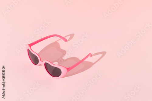 Pink sunglasses with sunlight shadow on pastel pink background. Minimal summer love concept.