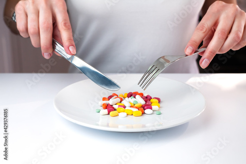 Female hands holds knife and fork and in front of her lie multicolored pills on a white plate. Taking vitamins and supplements. Close-up.