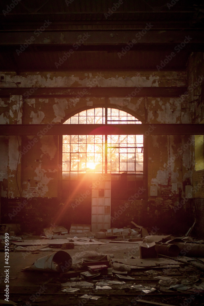A large semi-elliptical window through which the warm, afternoon sun sets inside to old abandoned brick and concrete warehouse with a lot of rubbish around.