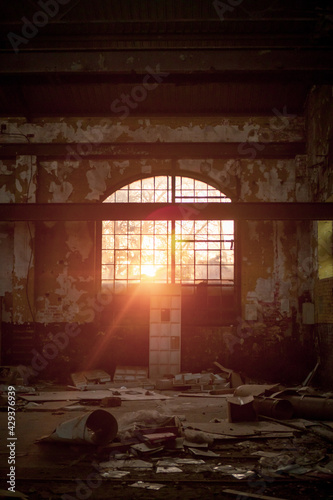 A large semi-elliptical window through which the warm  afternoon sun sets inside to old abandoned brick and concrete warehouse with a lot of rubbish around.