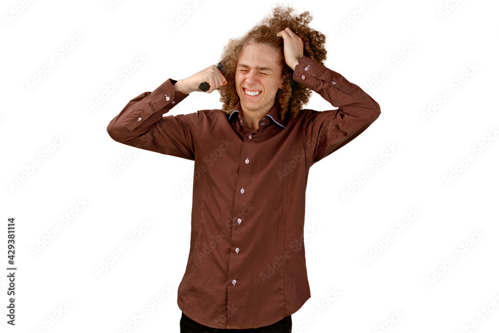 A long-haired curly-haired guy in a brown shirt on a white background uses a metal round comb. Emotions before a haircut in a hairdresser. Pain from combing A long-haired curly-haired guy in a brown shirt on a white background uses a metal round comb. Emotions before a haircut in a hairdresser. Pain from combing