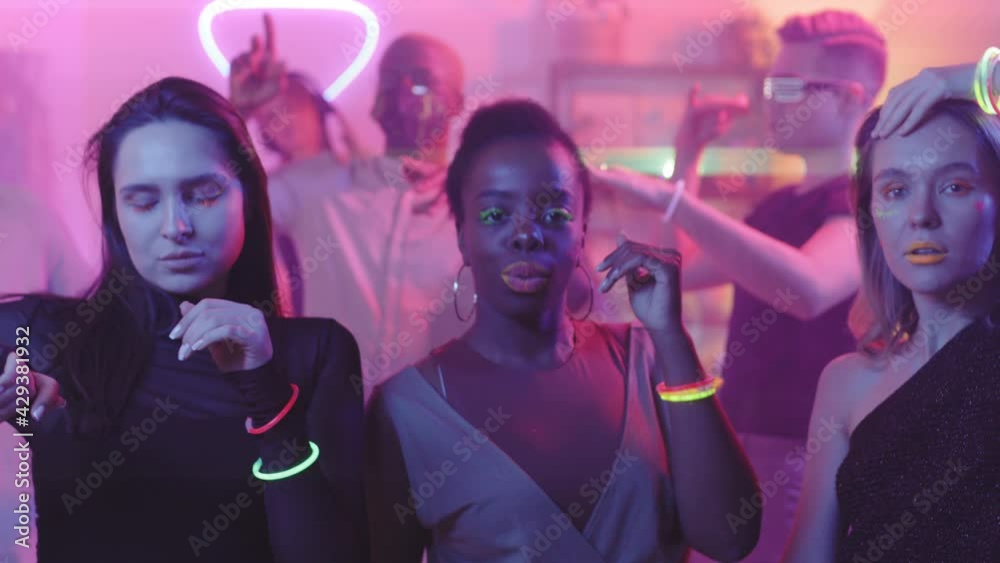 Medium shot of young women with vibrant makeup and neon bracelets looking at camera and dancing at house party with DJ Medium shot of young women with vibrant makeup and neon bracelets looking at camera and dancing at house party with DJ