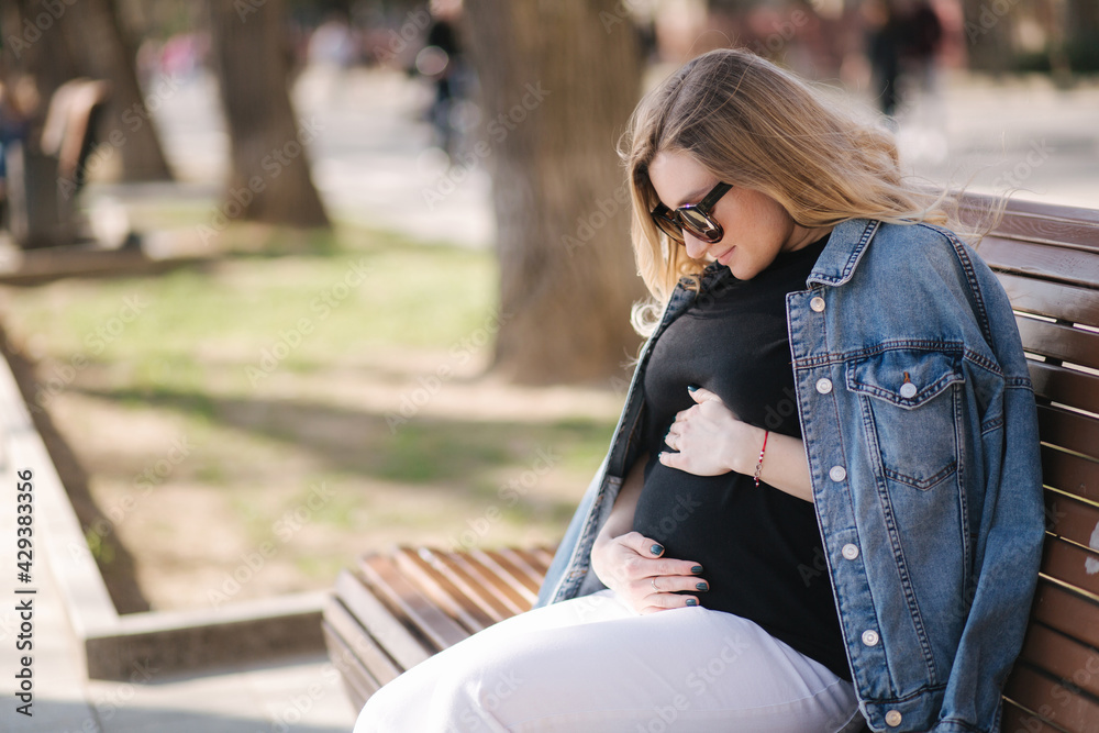 Happy pregnant woman put hands on belly and looking on tummy. Female sitting on the bench in park. Future mother breath fresh air Happy pregnant woman put hands on belly and looking on tummy. Female sitting on the bench in park. Future mother breath fresh air