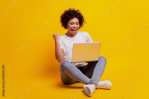 Smiling woman typing on laptop computer raise fist scream open mouth on yellow background