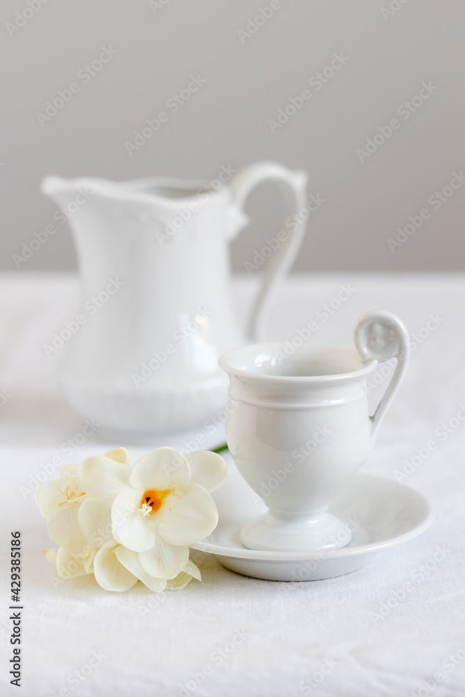 Minimalistic and clean coffee table styling. Elegant white cup of coffee with milk pitcher and blooms of freesia beside. Minimalistic and clean coffee table styling. Elegant white cup of coffee with milk pitcher and blooms of freesia beside.
