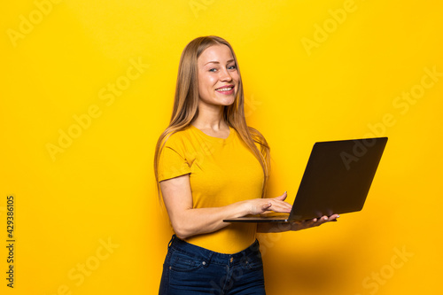 Portrait of a happy young blonde woman using laptop computer isolated over yellow background